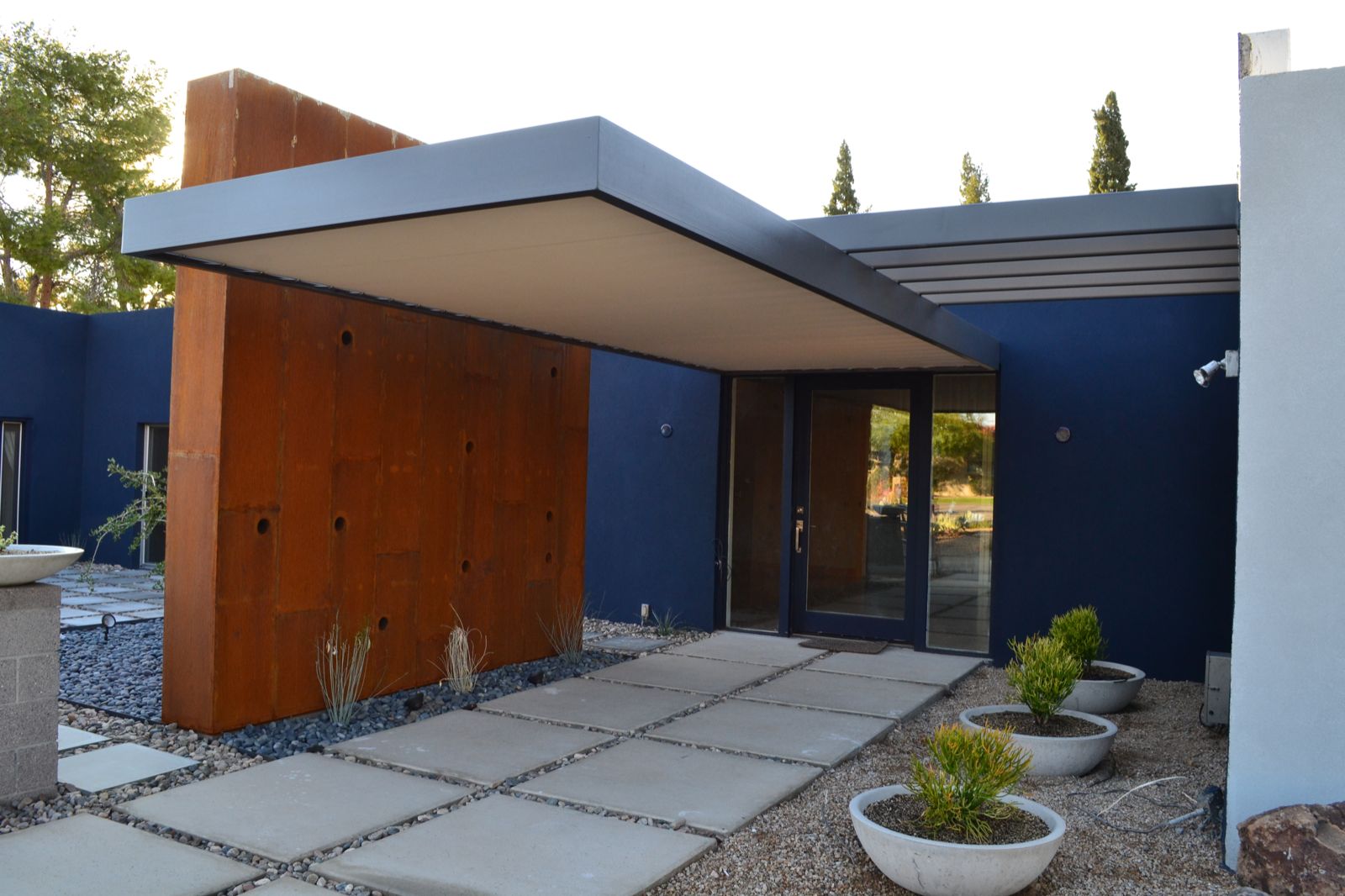 Picture of a hanging, grey, gray steel frame that is used to stretch a 20' by 8' shade cloth over the main entrance of a Scottsdale, AZ mansion, attached to a rusty, raw steel wall. Pavers and zero landscape that build the surroundings of a blue painted house wall.