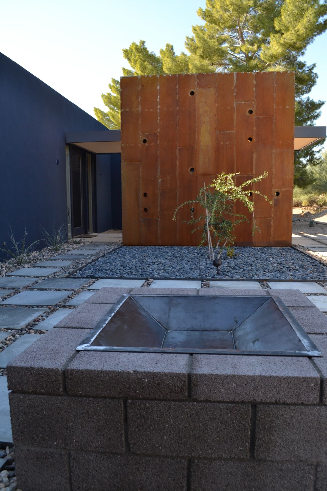 Picture of a square steel firepit in the foreground of a 12 foot by 12 foot tall raw, rusty steel wall.