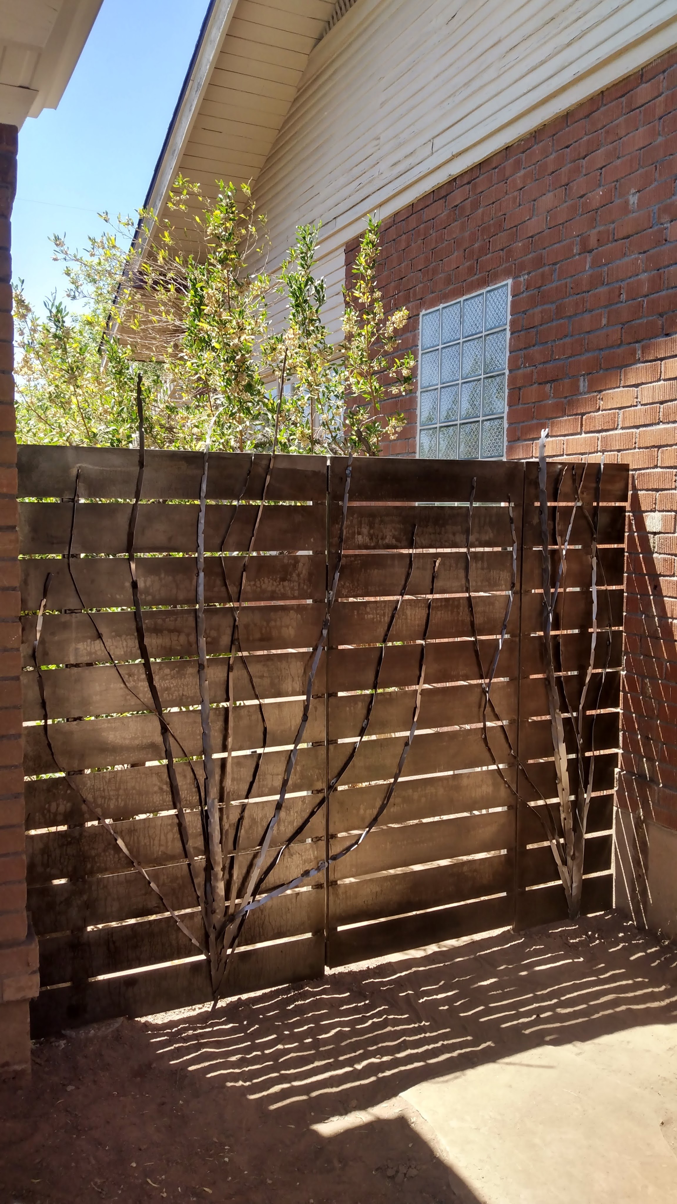 Picture of a modern steel fence that has ocotillo shaped steel forms welded to the front. Dusty ground next to brick-wall house in Coronado, AZ neighborhood