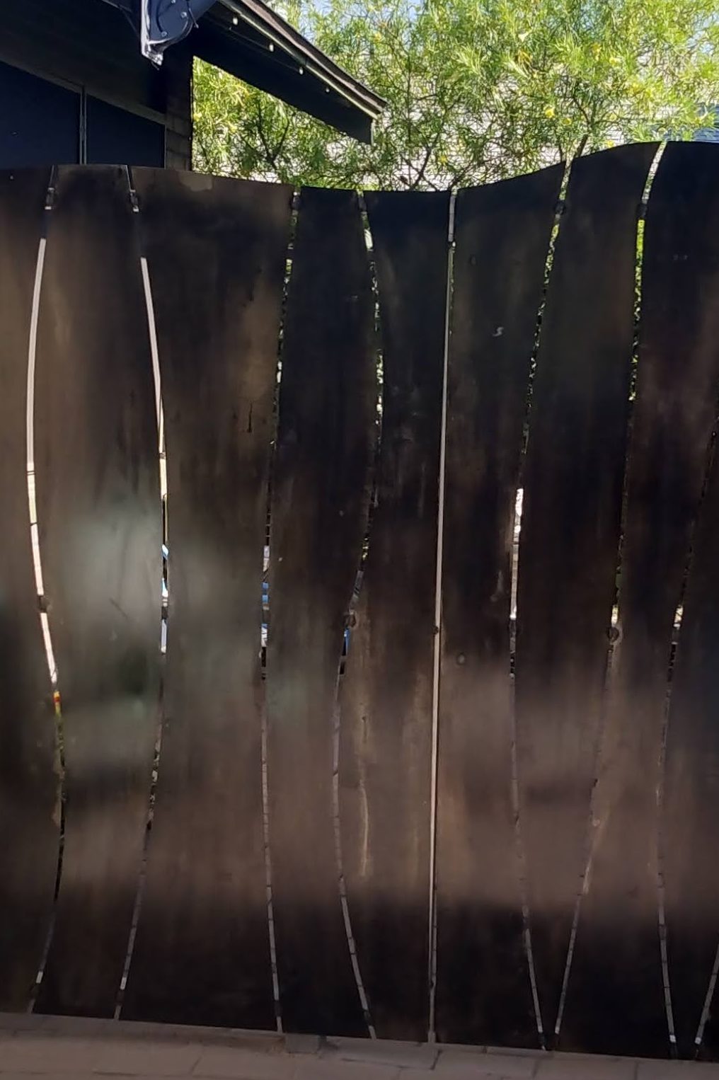 Detail of steel fence and gate with curved steel shaped sheet metal panels that are blackened with super blue bluing liquid. Trees and house roof in the background.